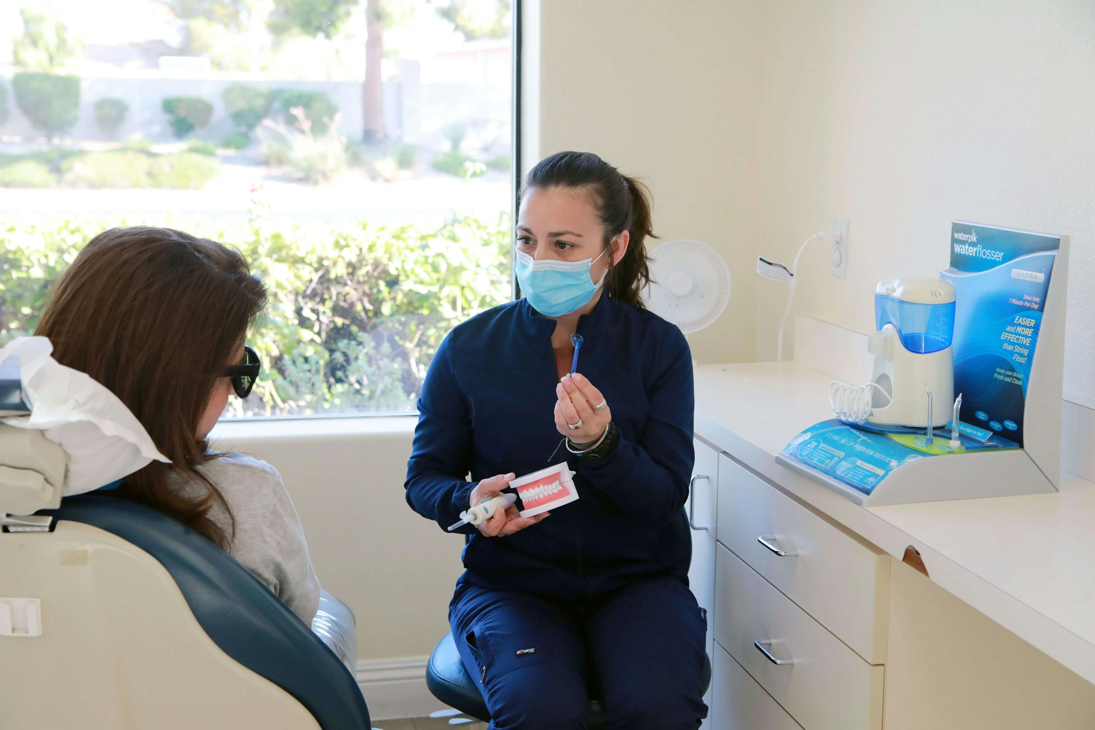 dental assistance in dark blue long sleave medical dress talks to a patient, explains how and why to use flossing procedure